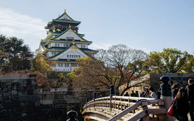 Osaka Castle's main tower rising above stone walls and a wooden bridge in autumn